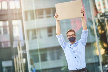 Young caucasian man, male activist wearing black protective mask holding empty sign board over the head while standing on city street. Protest during coronavirus outbreakの写真素材