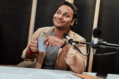 Portrait of young male radio host in headphones waking up, drinking coffee while getting ready for broadcasting in studioの写真素材