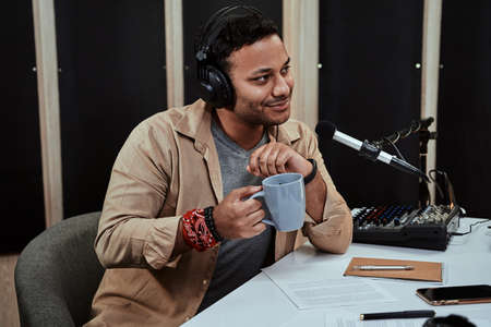 Portrait of young male radio host going live on air, talking with guest, holding a cup of coffee or tea while sitting in studioの写真素材