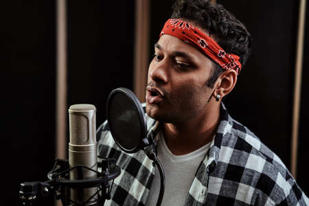 Portrait of young man, hip hop artist singing into a condenser microphone while recording a song in a professional studioの写真素材