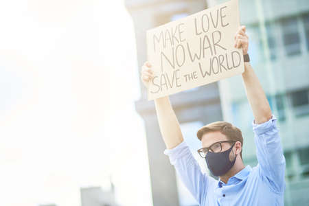 Antiwar demonstration. Young male activist wearing black protective mask holding banner or poster while protesting outdoorsの写真素材