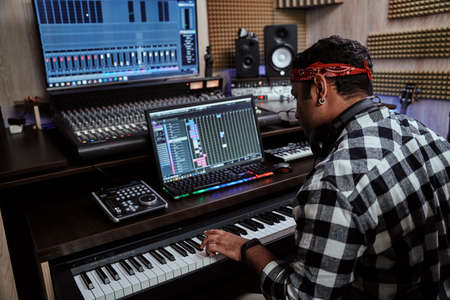 Young man, male artist looking focused while playing keyboard synthesizer, sitting in recording studioの写真素材