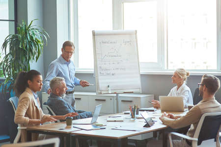 Start up team at brainstorming meeting. Confident mature businessman standing near flipchart in the modern office and discussing business goals with colleagueの写真素材
