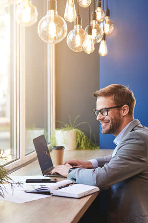 Happy freelancer. Side view of a young cheerful businessman using laptop and smiling while working remotely in cafeの写真素材