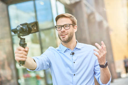 Young male vlogger wearing blue shirt and eyeglasses holding a gimbal with smartphone, talking and gesturing while recording new content for his blog outdoorsの写真素材