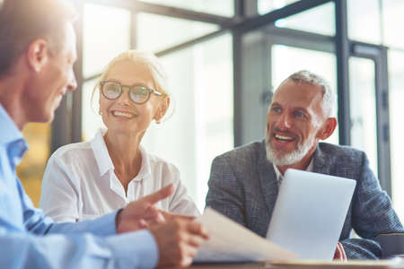 Successful happy business people discussing something and smiling while sitting at desk in the modern office or coworking spaceの写真素材