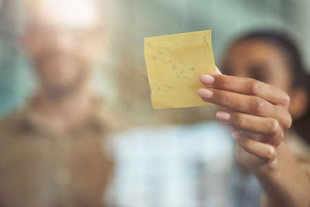 Young mixed race woman putting colorful sticky note on a glass wall while standing with her male collegue in the modern officeの写真素材