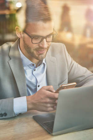 Chatting with friend. Vertical shot of a young handsome businessman or male freelancer using his smartphone while working online in cafe, laptop on wooden tableの写真素材
