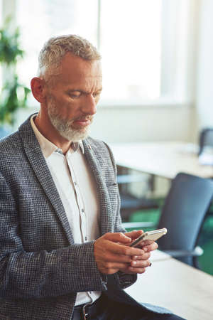 Confident mature businessman in formal wear using his smartphone while leaning on desk in the modern office or coworking space, vertical shotの写真素材