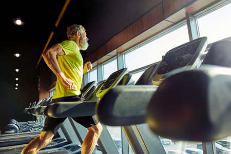 Side view of a athletic middle aged man in sportswear running on a treadmill in a gym, doing cardio...