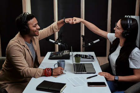 Portait of two cheerful radio hosts, young man and woman fist bump while talking, moderating a live show in studioの写真素材