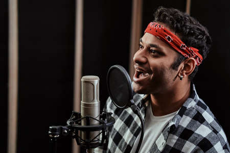 Portrait of young man, hip hop artist looking cheerful, singing into a condenser microphone while recording a song in a professional studioの写真素材