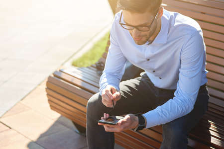 Working online outdoors. Young busy businessman wearing blue shirt and eyeglasses using smartphone while sitting on the bench on a sunny dayの写真素材