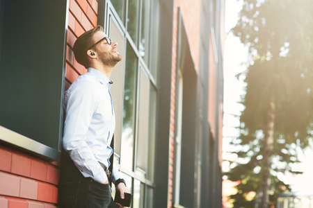 Side view of a young happy businessman listening to music with wireless earphones and mobile phone while leaning against brick wall outdoorsの写真素材