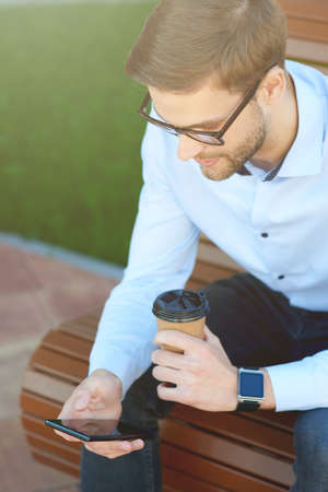 Vertical shot of a handsome businessman using smartphone and drinking coffee while relaxing on the bench outdoorsの写真素材