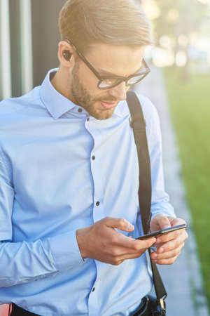 Making important call. Young handsome businessman wearing wireless earphones using smartphone while standing outdoorsの写真素材