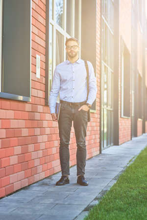 Full leghth of a young confident caucasian businessman wearing blue shirt and eyeglasses holding his laptop and looking at camera while standing outdoorsの写真素材