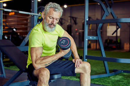 Bodybuilding in any age. Strong mature man in sportswear lifting heavy dumbbells and pumping his biceps while working out at gymの写真素材