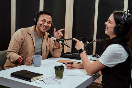 Portait of two cheerful radio hosts, man and woman smiling, pointing at each other, while moderating a live show in studioの写真素材