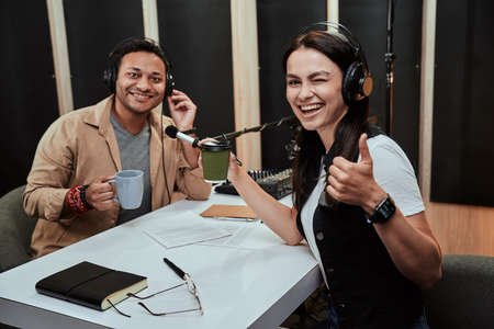 Portrait of two cheerful radio hosts, man and woman smiling at camera, drinking coffee or tea while getting ready for a live show in studio. Focus on female presenter showing thumbs upの写真素材