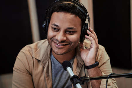 Close up of happy young male radio host smiling aside while broadcasting in studio, using microphone and headphonesの写真素材