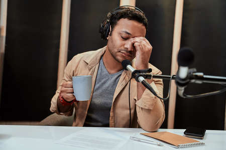Portrait of young male radio host in headphones looking tired, exhausted, drinking coffee while getting ready for broadcasting in studioの写真素材