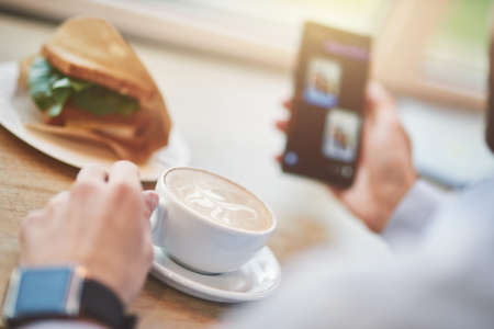 Cropped shot of a man drinking coffee and using smartphone while having breakfast in cafe