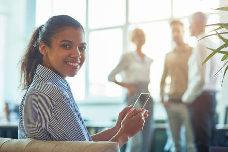 Young happy mixed race business woman sitting in the modern office, holding her smartphone and smiling at camera while her colleagues talking on the backgroundの写真素材