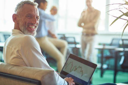 Young cheerful mature businessman sitting in the modern office, using laptop and smiling at camera while his colleagues talking on the backgroundの写真素材