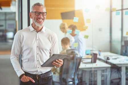 Happy mature businessman in classic wear holding digital tablet, looking at camera and smiling while...