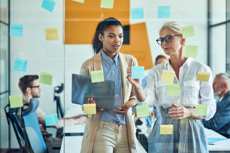 Brainstorming. Two happy female coworkers putting colorful sticky notes on a glass wall, sharing ideas and smiling while standing in the modern officeの写真素材