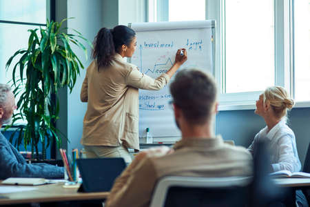Discussing business strategy. Young mixed race business woman drawing a graph on flipchart, giving a presentation to colleagues while having a meeting in the modern officeの写真素材