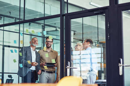 Group of creative coworkers standing in the modern office and looking at colorful sticky notes on a glass wall, discussing projectの写真素材