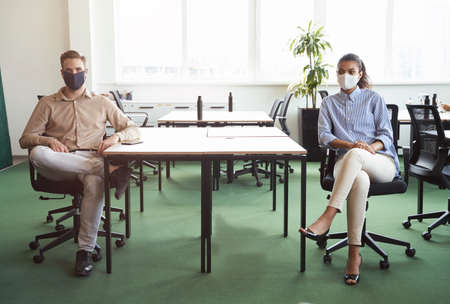 Two young diverse colleagues wearing protective face mask looking at camera while sitting at desk in...