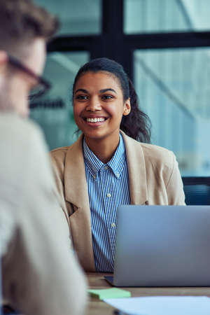 Vertical shot of a young cheerful mixed race female office worker working on laptop in the modern office, talking with colleague and smilingの写真素材
