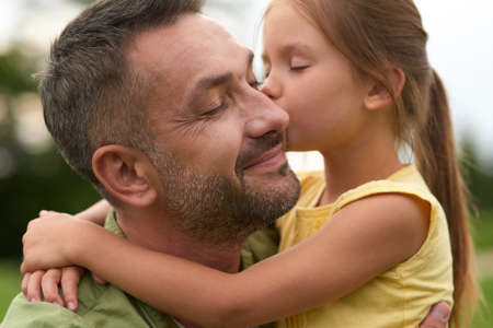 Cute little girl hugging and kissing her happy dad while spending time together outdoors on a warm dayの写真素材