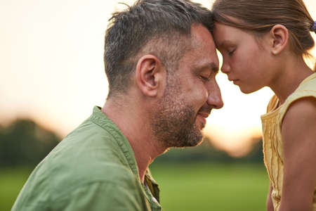 Close up shot of young father and adorable little daughter touching foreheads, standing with eyes closed while spending time outdoors in the park on a summer dayの写真素材