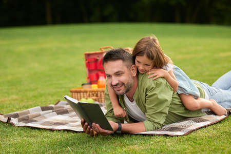 Curious little girl distracting, playing with her dad when he reads a book while lying on a blanket. Happy father and his daughter having a picnic in the parkの写真素材