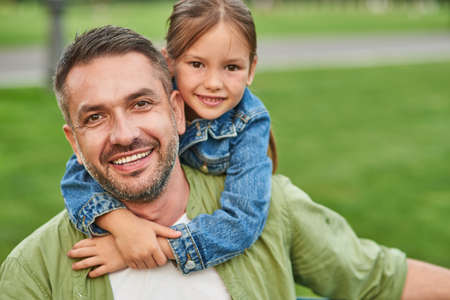 Portrait of young joyful father and his cute little daughter looking at camera and hugging while spending time together outdoors, sitting on a green grassの写真素材