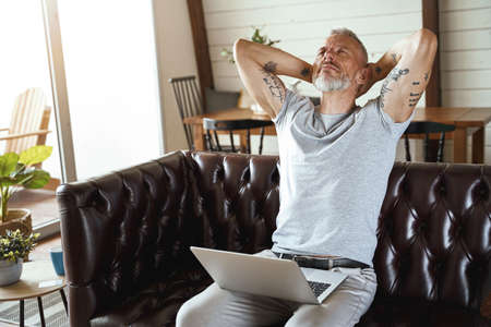 In search of inspiration. Thoughtful mature man in casual clothes looking upside and thinking about business while sitting on sofa in the living room, working on laptopの写真素材