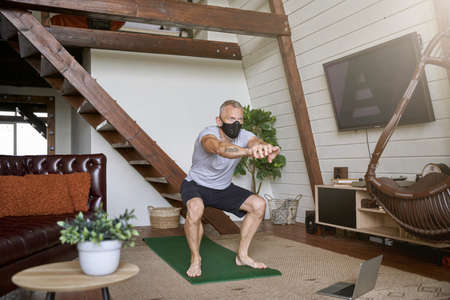 Staying active in self isolation. Middle aged athletic man wearing face protective mask exercising at home while watching online workout on laptopの写真素材