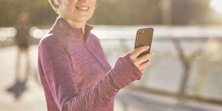 Cropped portrait of joyful active mature woman in sportswear using her smartphone, checking results after jogging outdoors on a sunny dayの写真素材