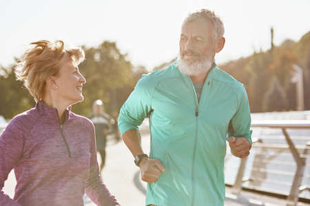 Keep moving. Cheerful active mature family couple in sportswear looking at each other, having a talk while jogging together on a sunny day. Joyful senior couple doing sport outdoorsの写真素材