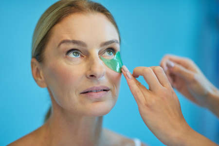 Beauty procedures. Beautiful mature lady looking upside while female beautician putting patches under her eyesの写真素材