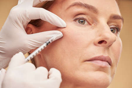 Beautician hands in gloves holding syringe near female cheekbone