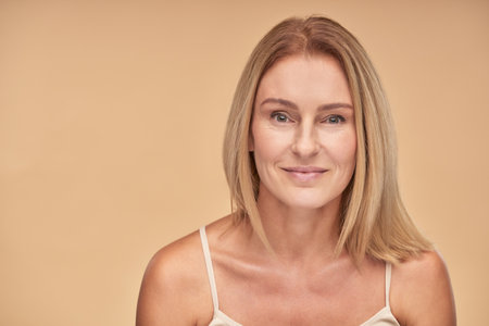 Natural beauty. Portrait of a charming middle aged caucasian woman with clean skin looking at camera while posing over beige background in studioの写真素材