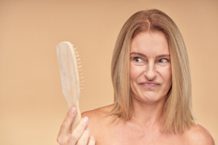 Hair loss problem. Frustrated and upset mature blonde woman looking at combの写真素材