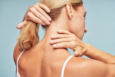 Rear view of a mature woman touching her neck with soft and clean skin while standing against blue backgroundの写真素材