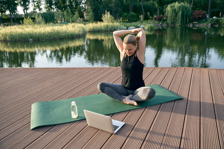Beginner. Sporty beautiful woman exercising, watching instructional videos on a laptop while doing yoga on a mat in nature on a sunny dayの写真素材