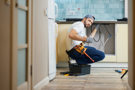 The work is done. Young repairman, professional plumber wearing tool belt looking at camera and showing thumbs up while crouching on the floor, fixing a pipe in the kitchen indoorsの写真素材